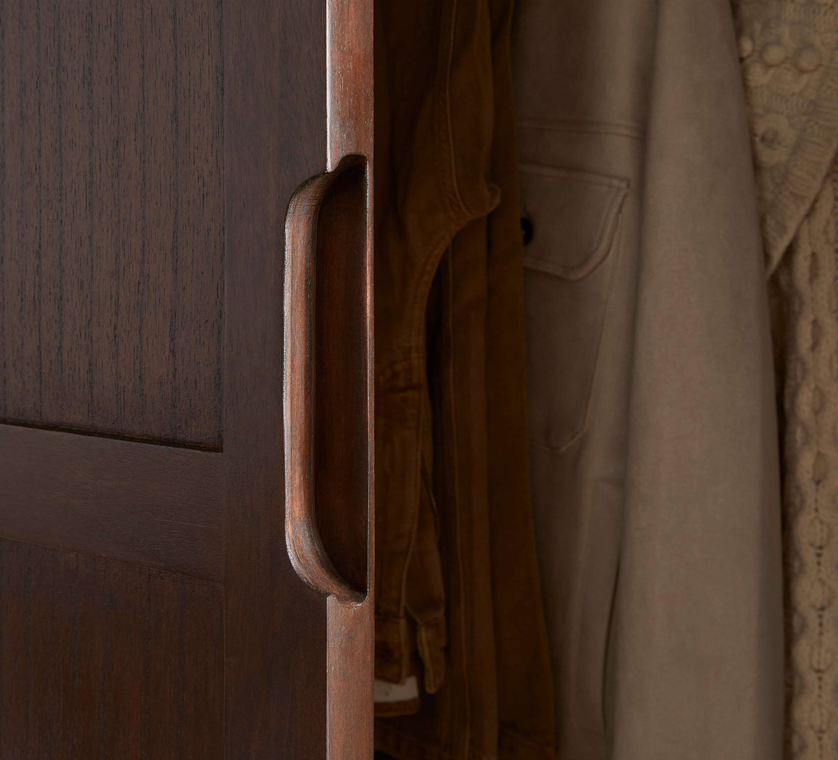 Close-up of the carved handle on the Fishe & Lilly Norway Dark Walnut Wardrobe with hanging clothes.