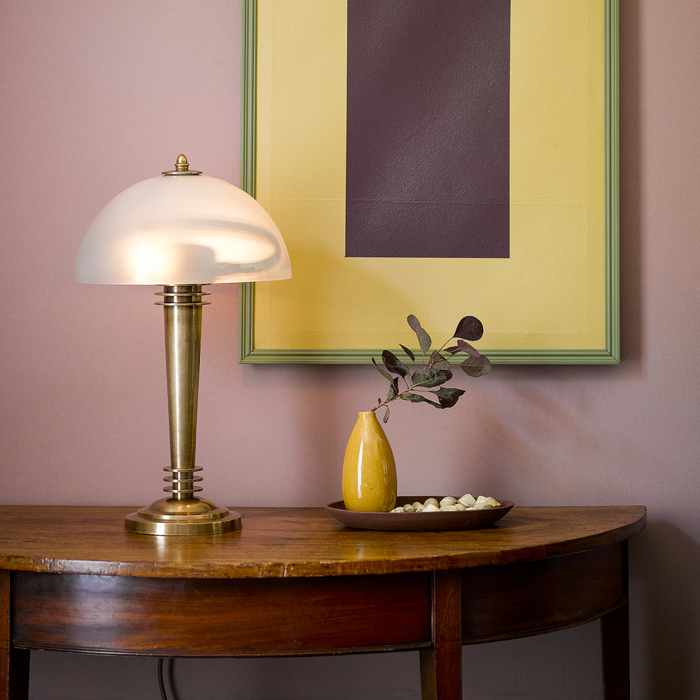 Pooky - Pietro Table Lamp - Brass and Glass, an Art Deco style lamp with a frosted dome shade, glowing on a wooden table.