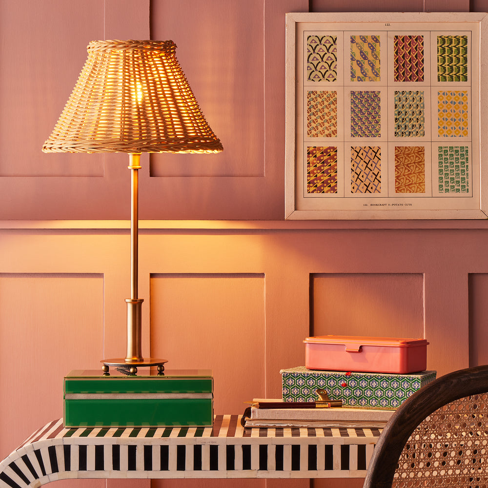 Pooky - Regular Pod Table Lamp Base - Antique Brass paired with a wicker shade on a striped desk.