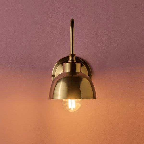 Pooky - Carmel Wall Light - Antiqued Brass Finish showcasing a stylish brass wall arm with illuminated bulb on a pink background.