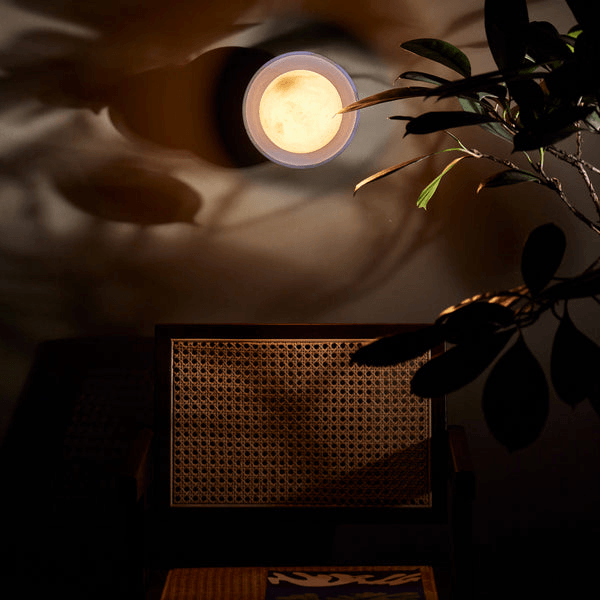 Pooky - Luna Wall Light - Klein Blue illuminating a wall above a cozy chair with a plant shadow.