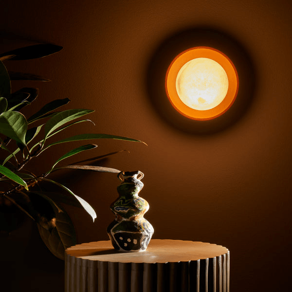 Pooky - Luna Wall Light - Terracotta Finish illuminating a wall beside a decorative vase and plant.