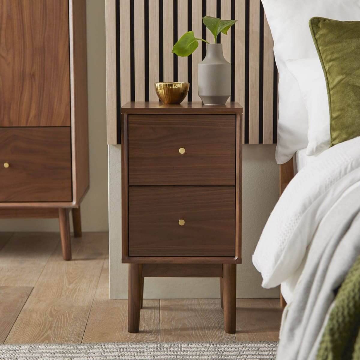Fishe & Lilly Wright Narrow Walnut Bedside Table with two drawers and solid wood legs beside a bed.