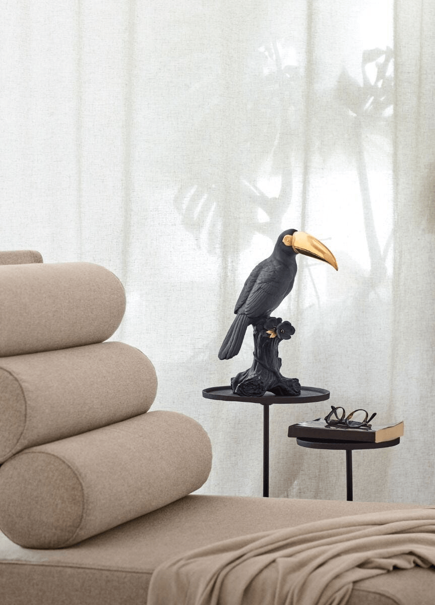Lladró - Toucan Sculpture - Black-gold - Limited Edition displayed beside a cozy chair and accessories.