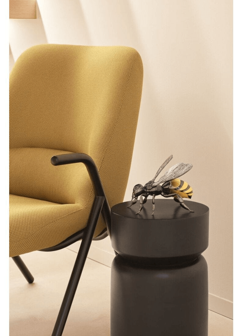 Lladró - Bee Sculpture on a black pedestal next to a stylish yellow chair, showcasing exquisite porcelain craftsmanship.