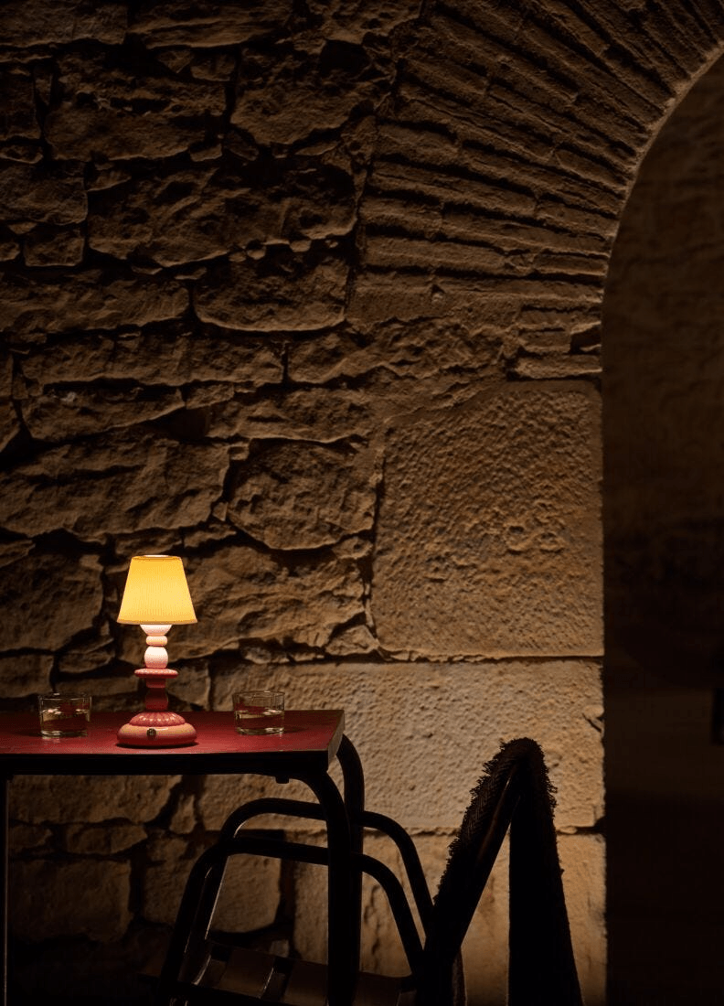 Lladró - Lotus Firefly Table Lamp - Coral illuminating a table with stone wall background, showcasing its porcelain design.