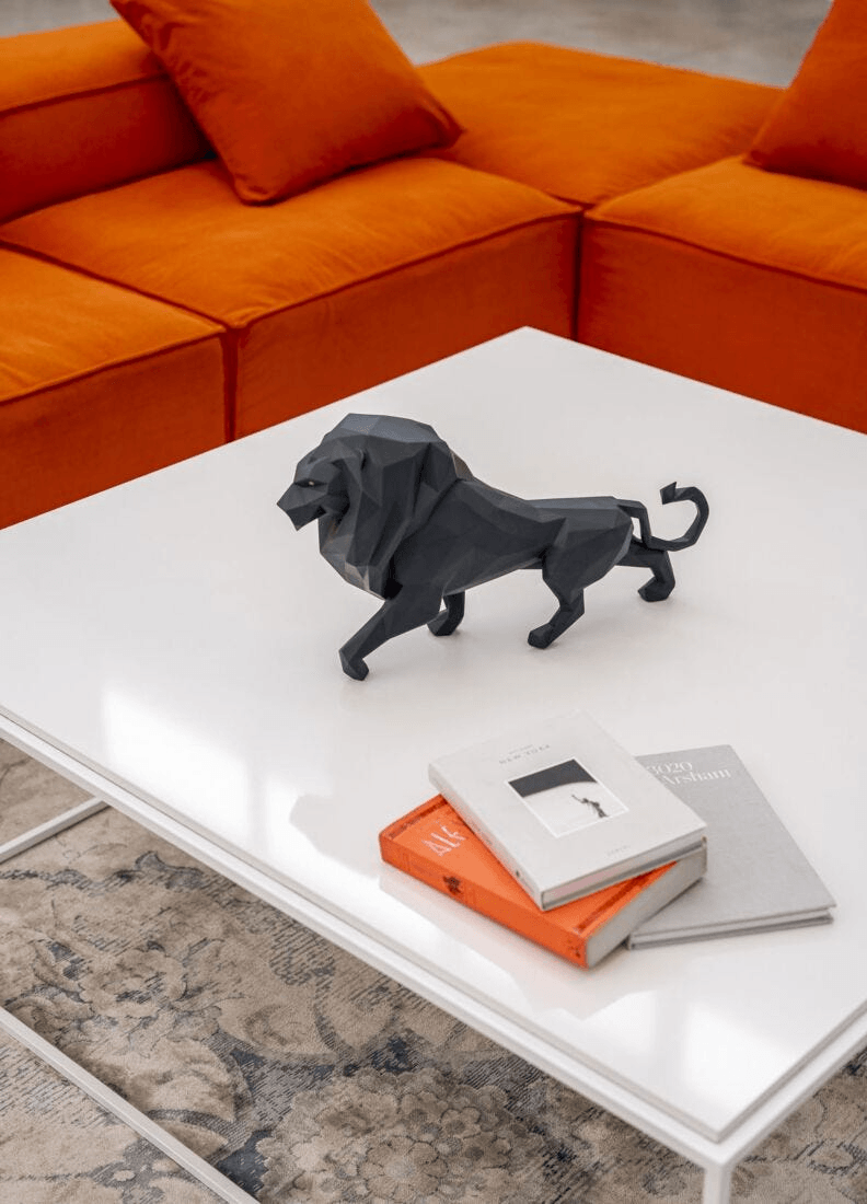 Lladró - Lion Sculpture - Matte Black displayed on a white table with books and orange couch in the background.