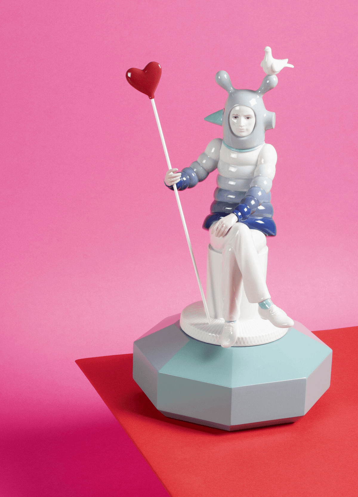 Lladró - The Lover I Sculpture, a porcelain figure by Jaime Hayon with a heart and dove, set against a pink background.