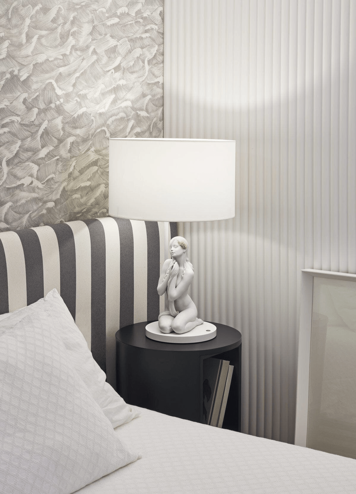 Lladró - Inner Peace Table lamp made of white Lladro Porcelain on a black nightstand next to a bed.