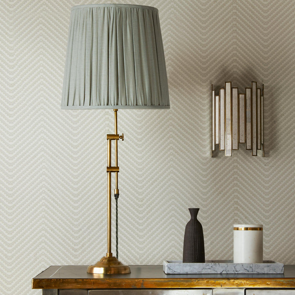 Pooky - Slider Table Lamp Base - Brass with a grey pleated shade on a table against patterned wallpaper.