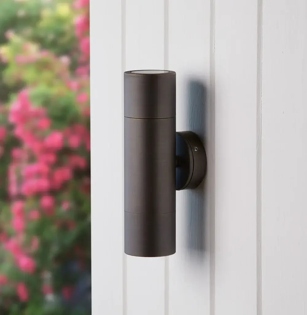 Pooky Portreath IP65 Up and Down Pillar wall light in Bronze installed on a white wall with blurred greenery in the background.
