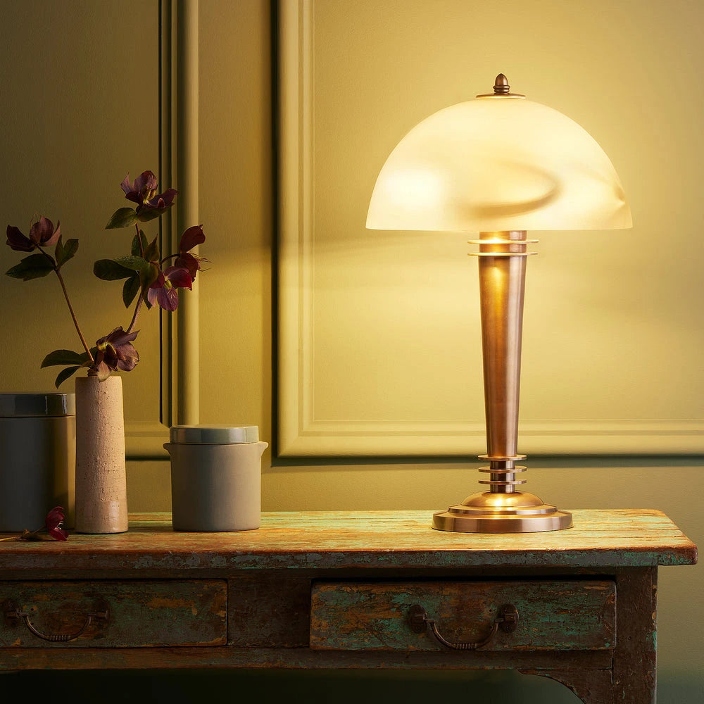Art Deco Pooky - Pietro Table Lamp - Brass and Glass lit on a rustic wooden table.