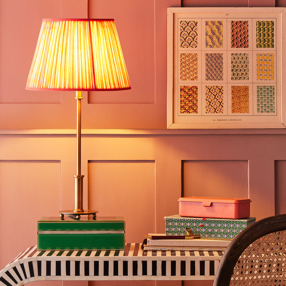 Pooky - Regular Pod Table Lamp Base - Antique Brass shown lit with a pleated shade on a striped desk.